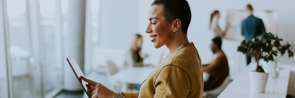 A smiling person using a tablet in a bright office environment with colleagues in the background.