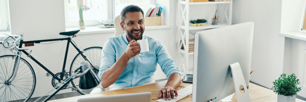 Man with a coffee mug smiling while working at a computer in a bright office setting.